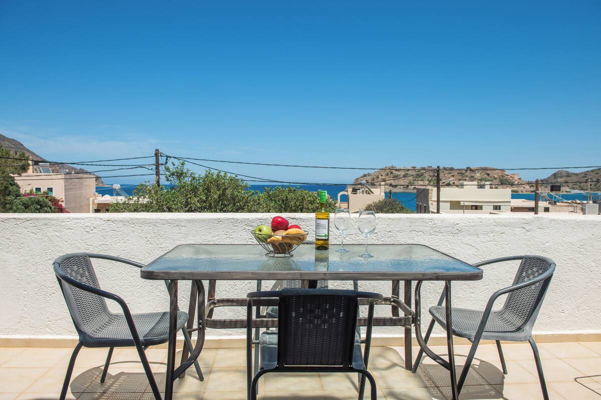 Ein Tisch mit Glasplatte, zwei Stühle, eine Obstschale und eine Flasche Olivenöl auf einer Terrasse mit Meerblick.