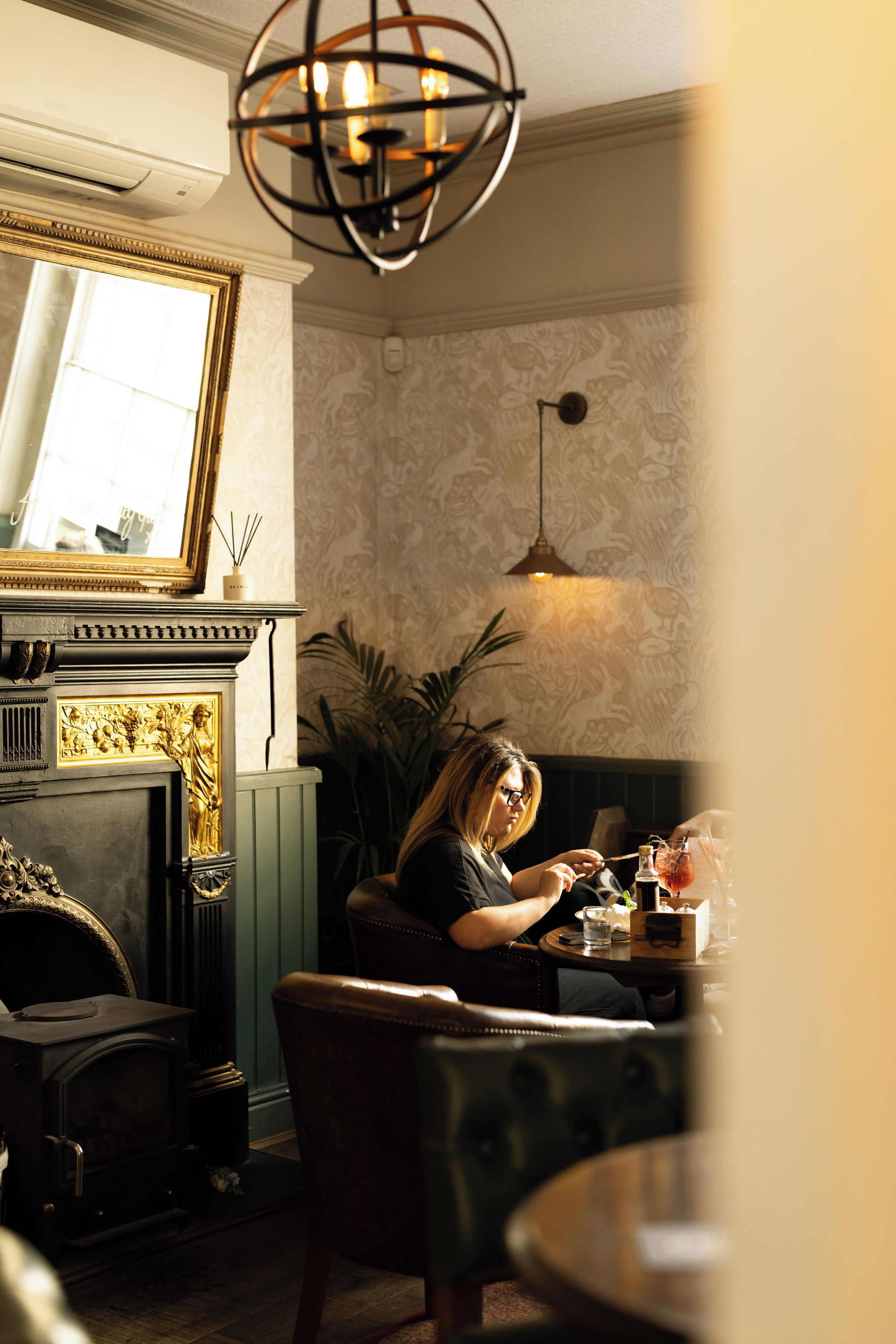 A room with a fireplace, a mirror, a chandelier, and a table with a drink.