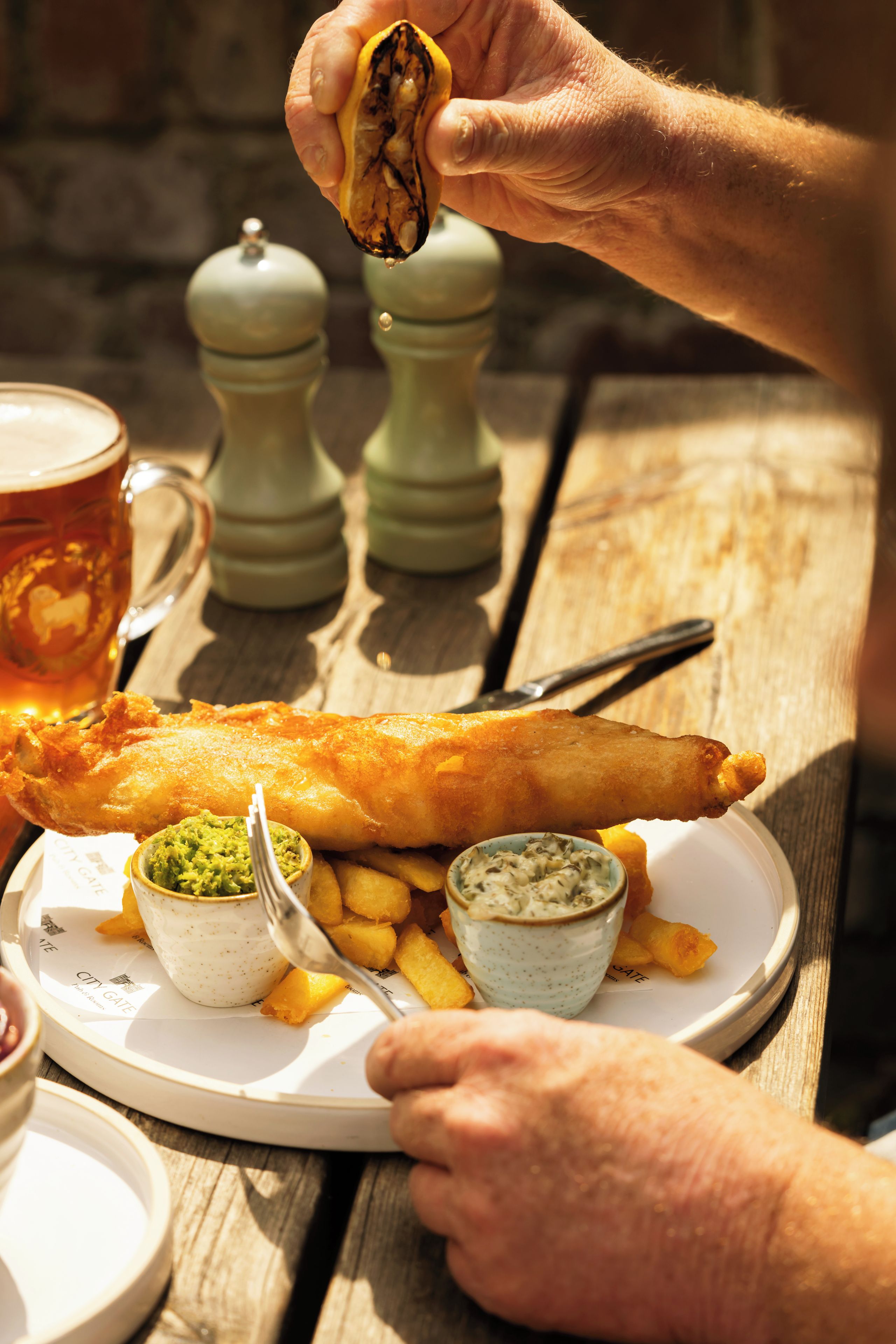 A meal consisting of a large fish and chips dish with two small bowls of sauce, served on a white plate with a wooden table.