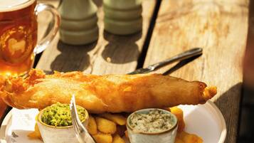 A meal consisting of a large fish and chips dish with two small bowls of sauce, served on a white plate with a wooden table.