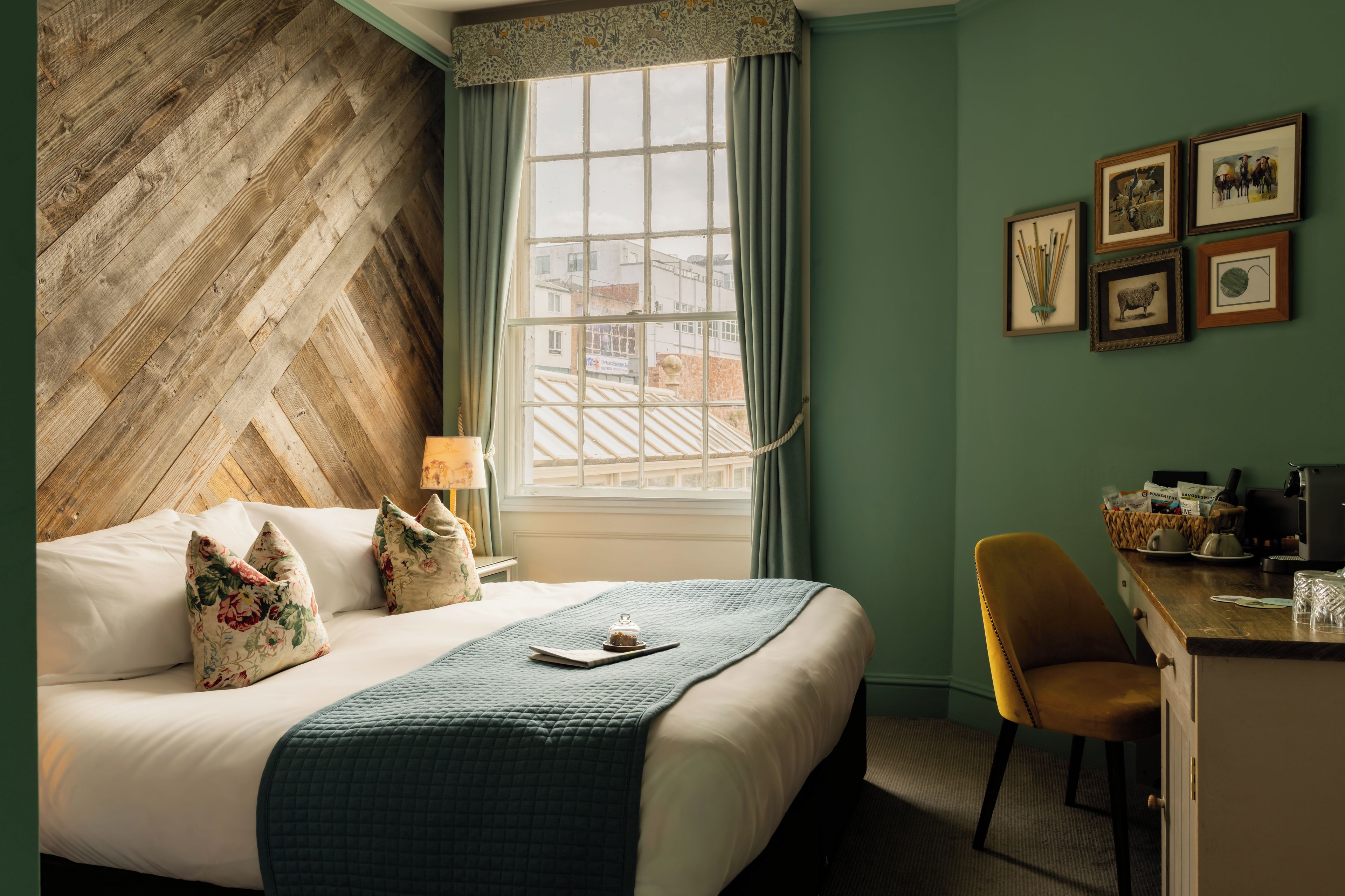 Habitación de hotel con cabecera de diseño greca, cama con almohadas florales, escritorio con cafetera y cuadros en la pared.