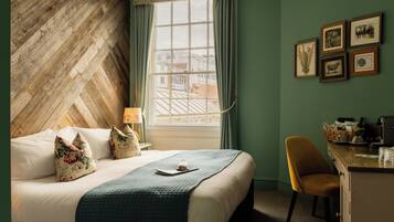 Habitación de hotel con cabecera de diseño greca, cama con almohadas florales, escritorio con cafetera y cuadros en la pared.