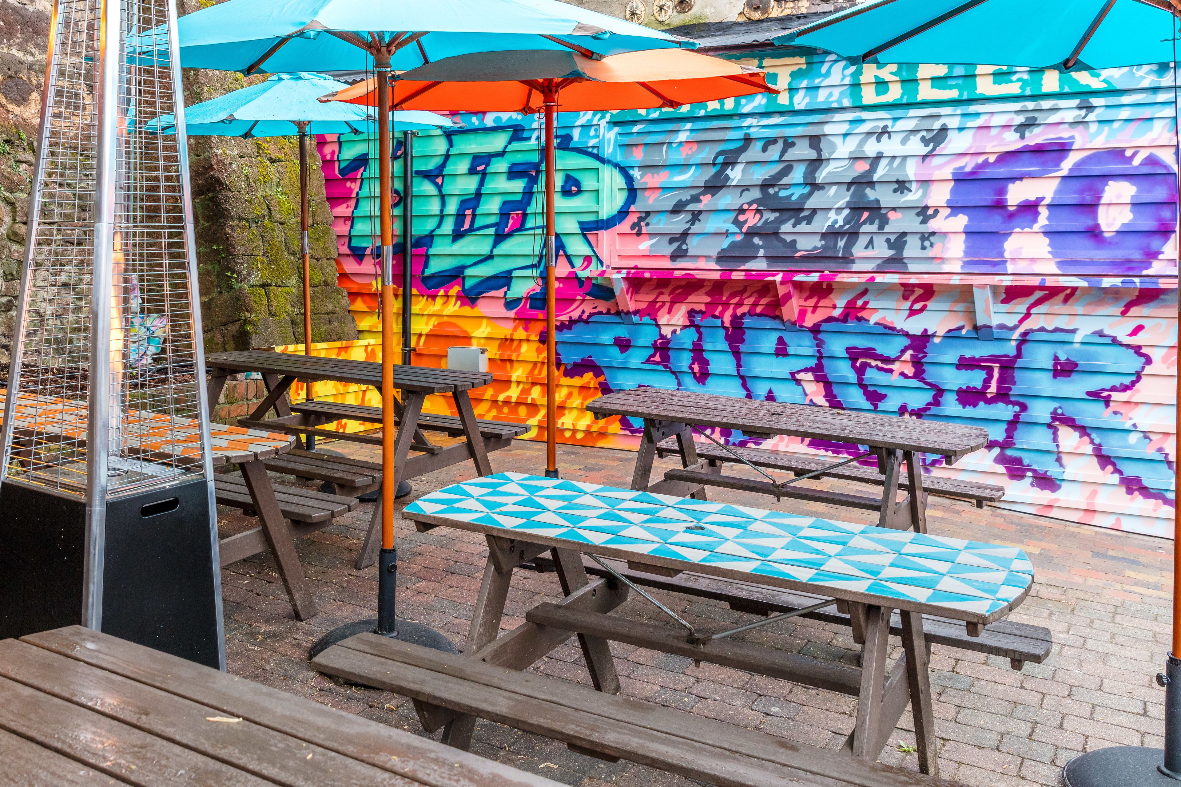 Ein Außenbereich mit Picknicktischen und Sonnenschirmen, vor einer Wand mit bunten Graffiti.