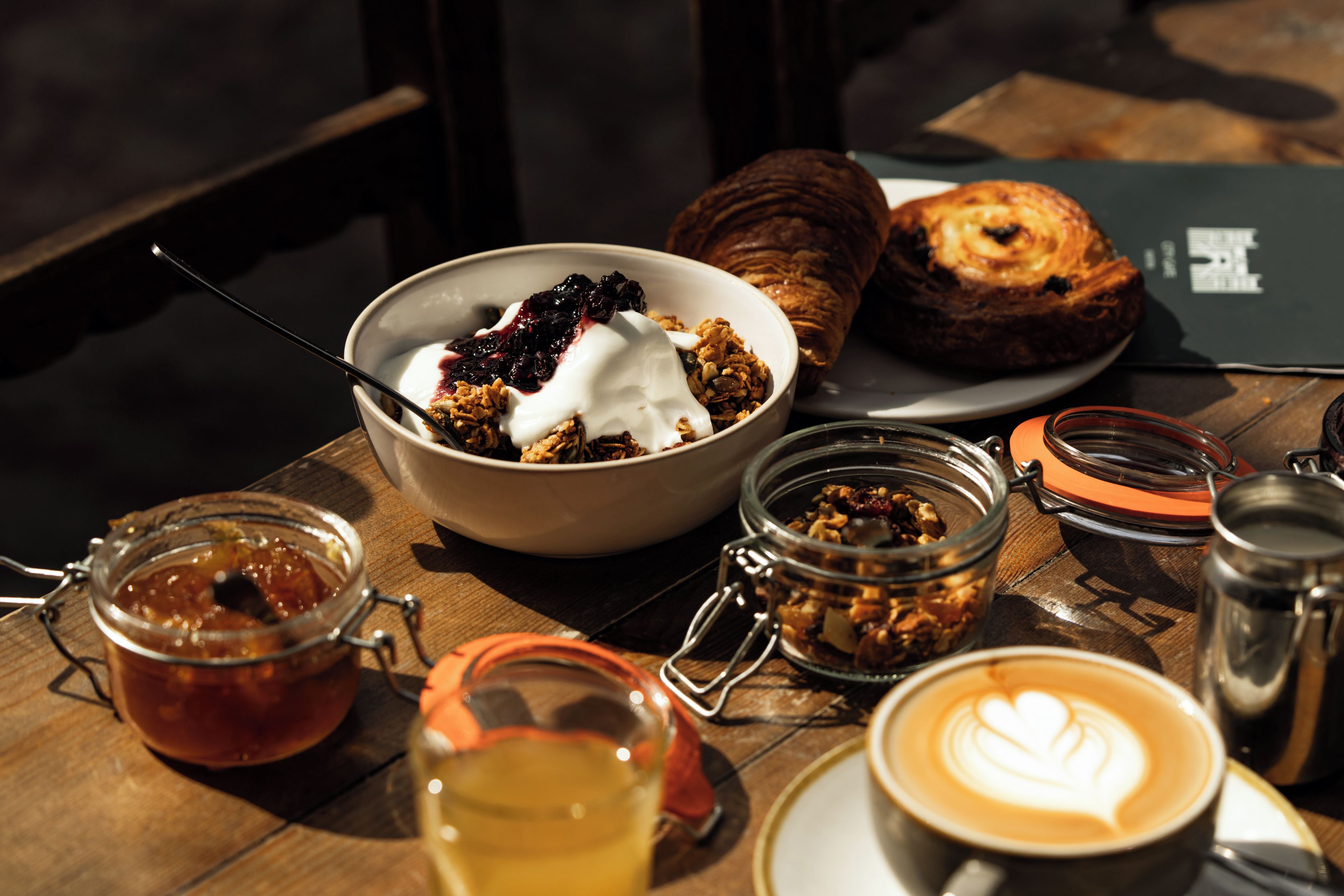 Une table en bois dressée pour le petit-déjeuner, avec un bol de céréales, un croissant, une tasse de café décorée de motifs en latte art et un pot de confiture.