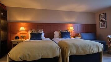 Quarto de hotel com duas camas, cada uma com cabeceira, mesas de cabeceira, luminárias e uma cadeira com padrão floral.