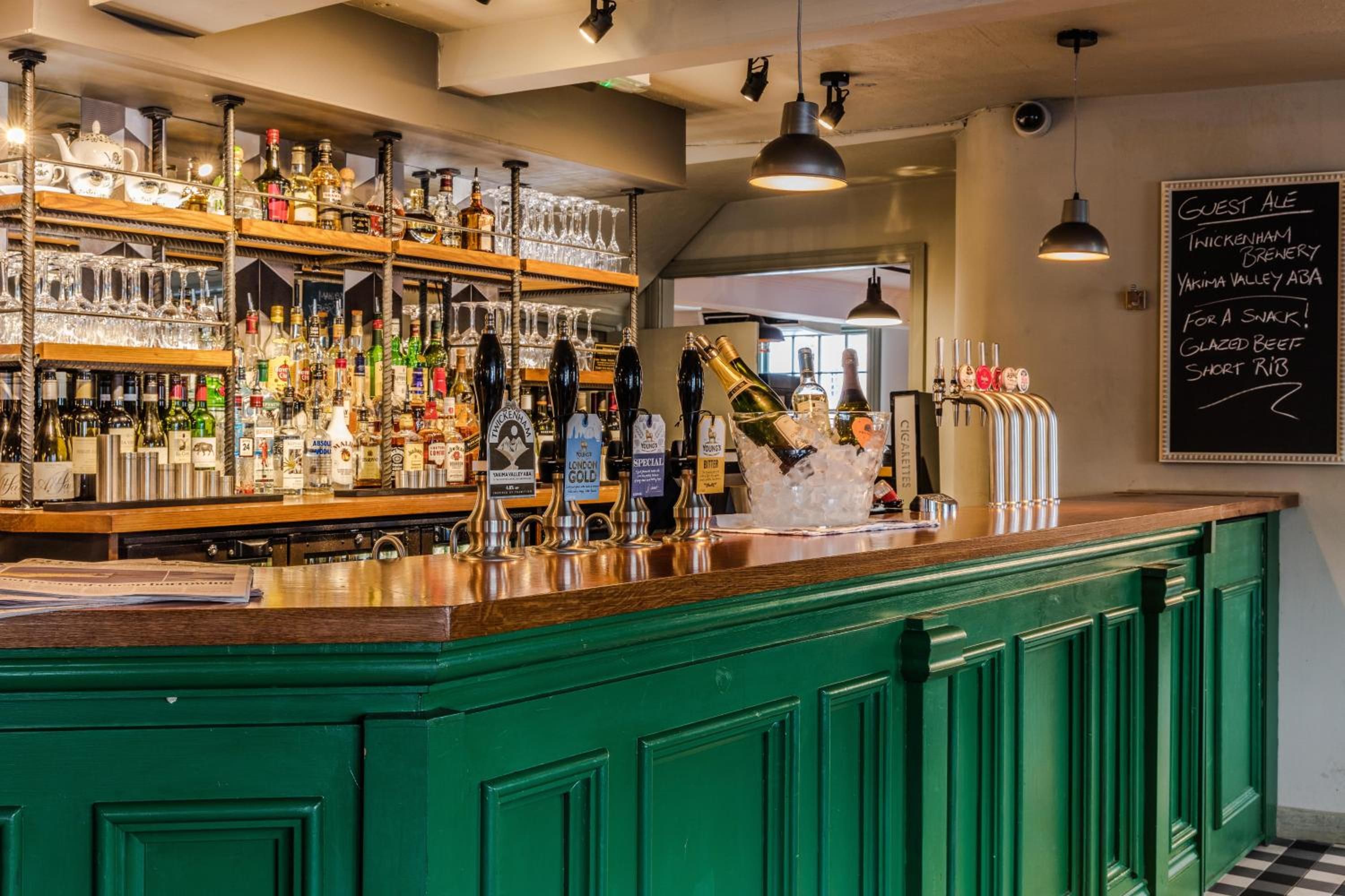 Un espace bar avec un comptoir vert, diverses bouteilles, des verres et un menu écrit à la craie sur un tableau noir.