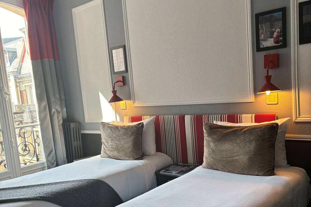 A hotel room with two beds, a large window with curtains, a wall-mounted lamp, and a framed picture.