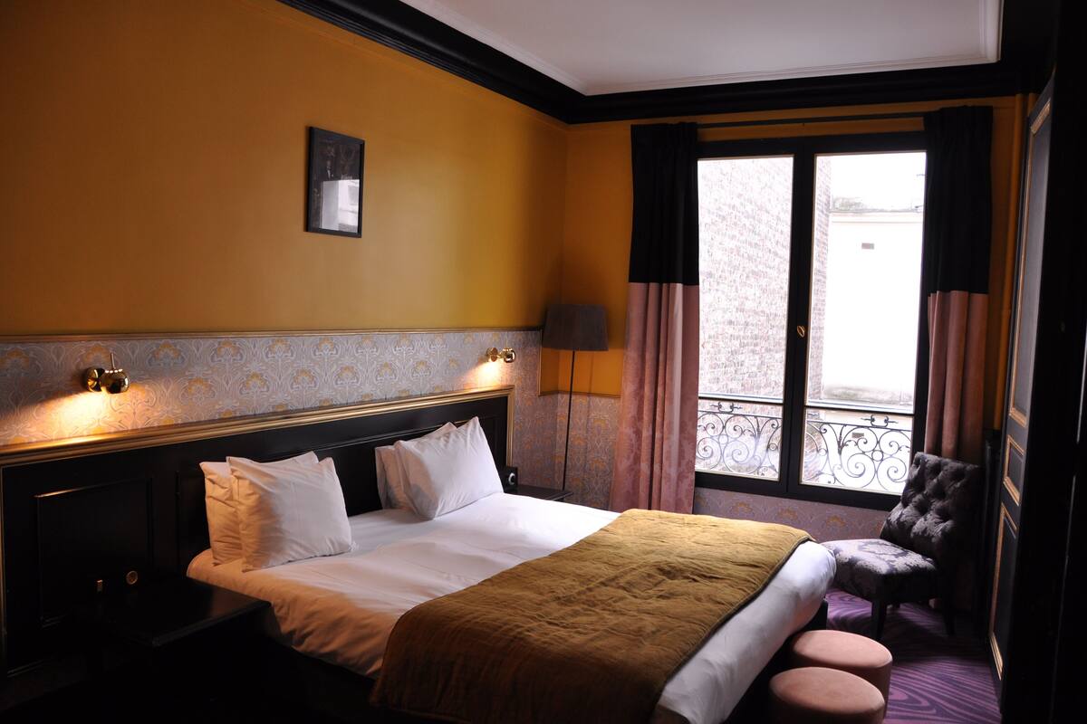 A hotel room with a large bed, a chair, a window with a view, and a lamp.
