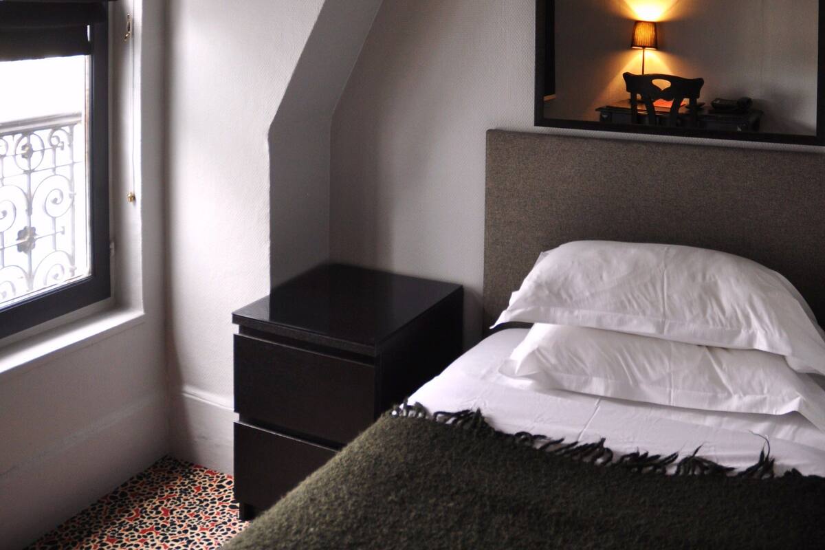 A bedroom with a sloped ceiling, a bed with white bedding, a nightstand, a lamp, a TV mounted on the wall, and a window with a view of a balcony.