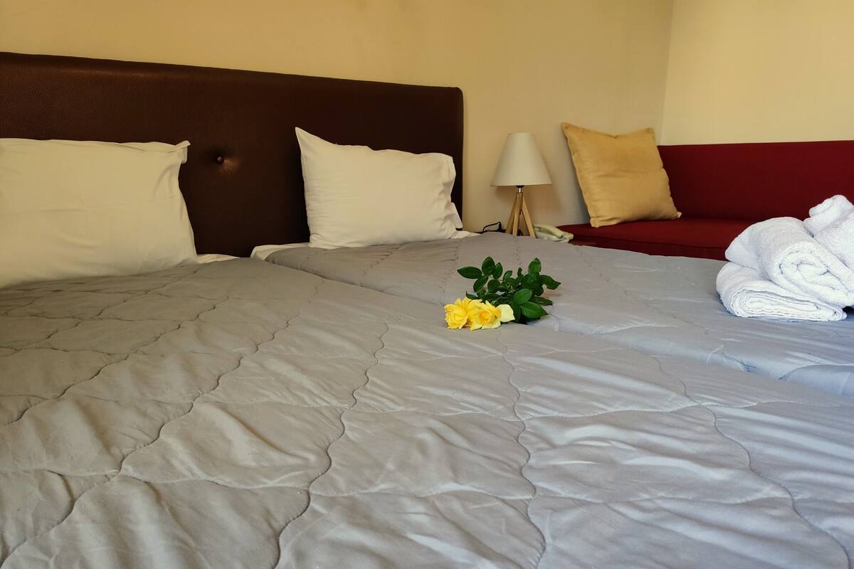 A neatly made bed with a grey quilt, two white pillows, and a small bouquet of yellow flowers.
