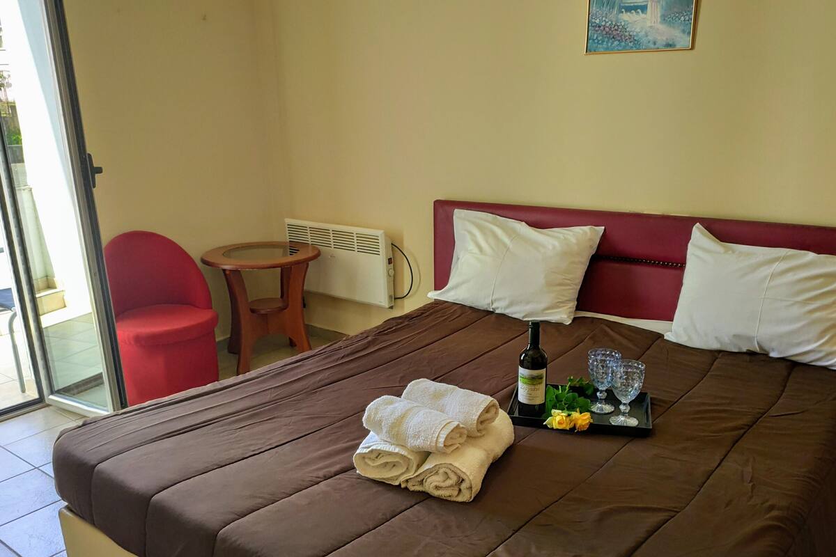 A neatly made bed with a brown bedspread, two white pillows, a bottle of wine, and a tray with glasses and flowers.