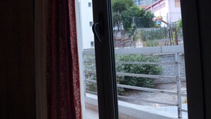 A window with a view of a building with a red roof, a metal fence, and a large rock formation in the background. - King Lodge (Kalabaka)