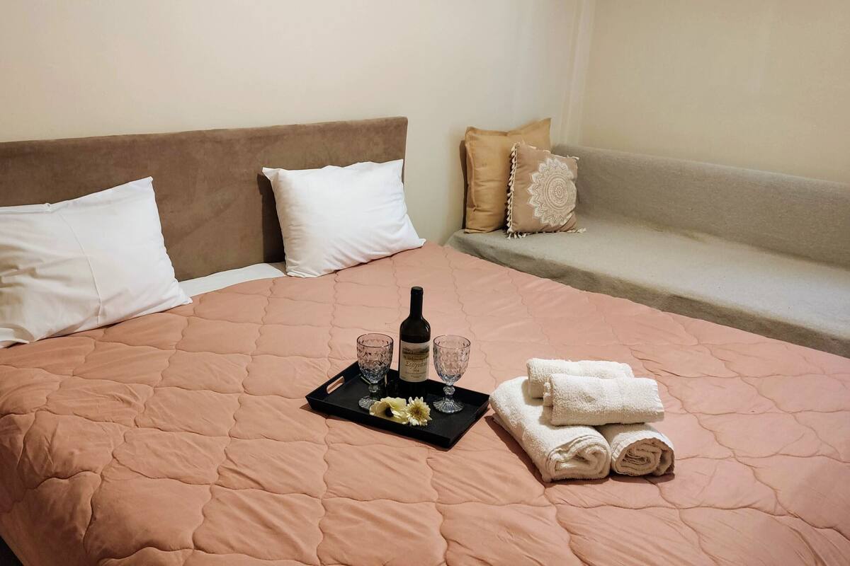 A neatly made bed with a pink quilt, a tray with a bottle, glasses, and towels.