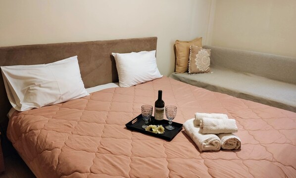 A neatly made bed with a pink quilt, a tray with a bottle, glasses, and towels.