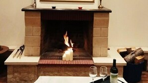 A fireplace with a red panel above it, a table with glasses and a bottle, and a log fire burning. - King Lodge (Kalabaka)