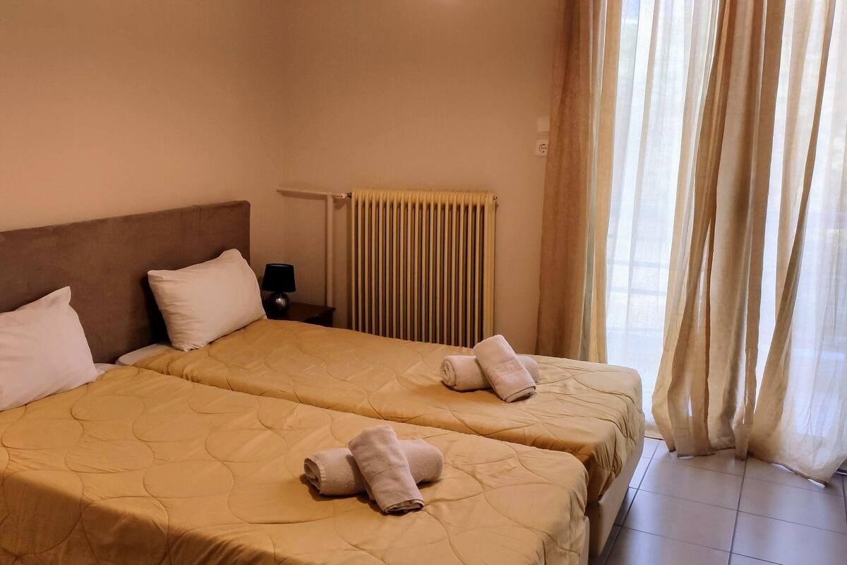 A hotel room with two beds, beige bedding, a radiator, and a window with curtains.
