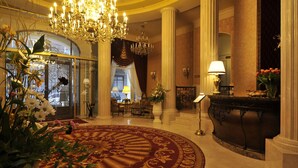 A grand lobby with a large chandelier, columns, a red carpet, and a reception desk with a vase of flowers.