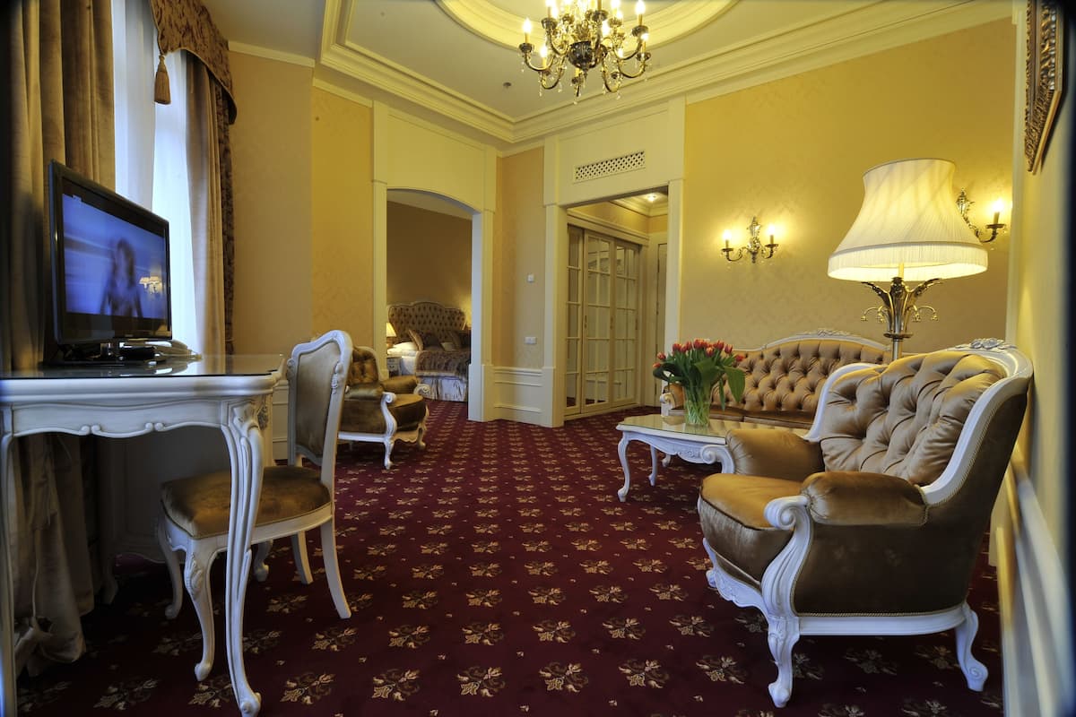 A hotel room with a red carpet, a television, a sofa, a chair, a table with a lamp, and a door leading to another room.