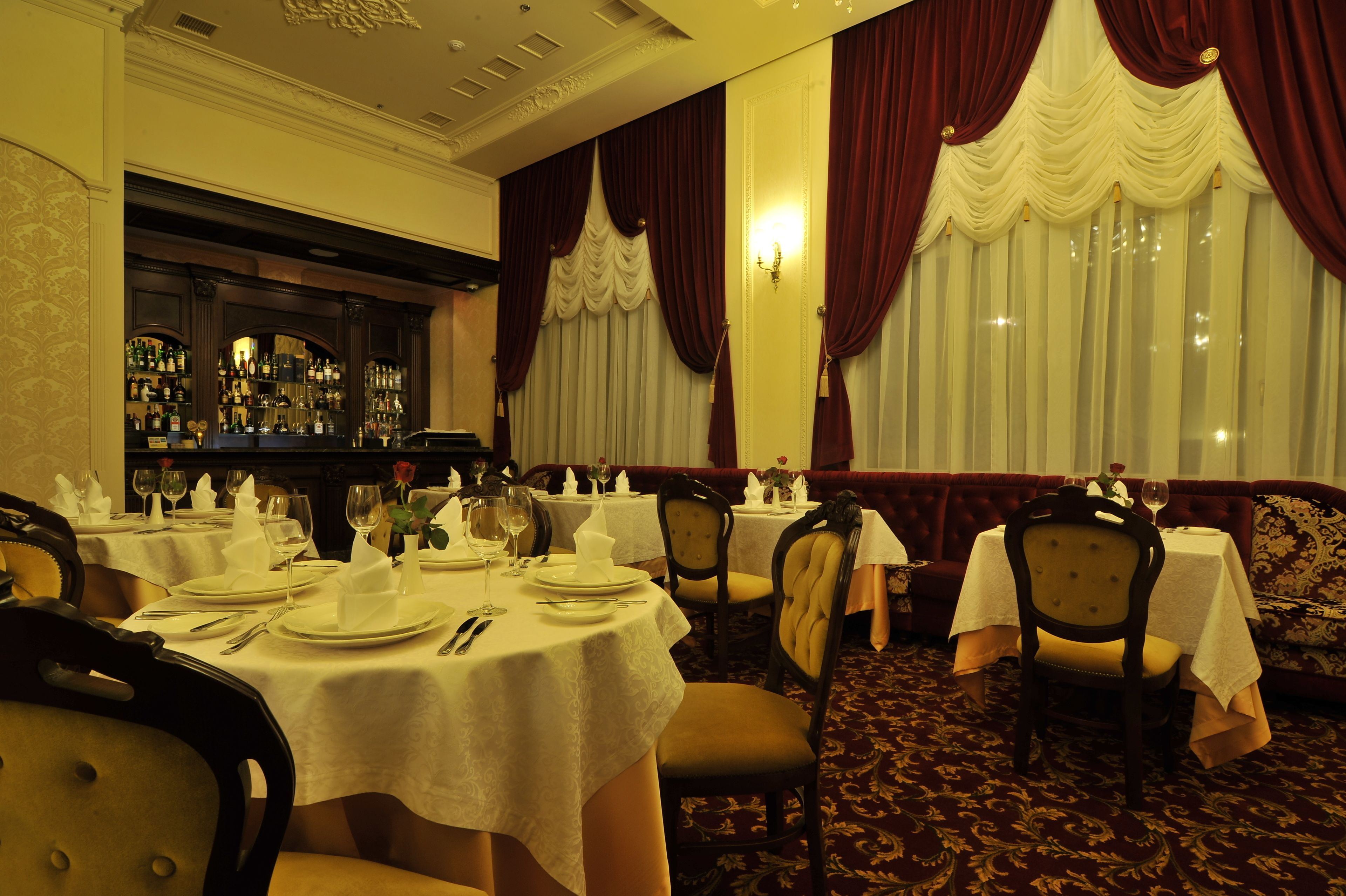 Un restaurant avec des tables rondes dressées pour le service, un bar avec des bouteilles exposées et des rideaux rouges.