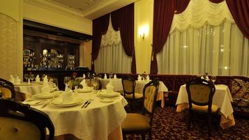 Un restaurant avec des tables rondes dressées pour le service, un bar avec des bouteilles exposées et des rideaux rouges.
