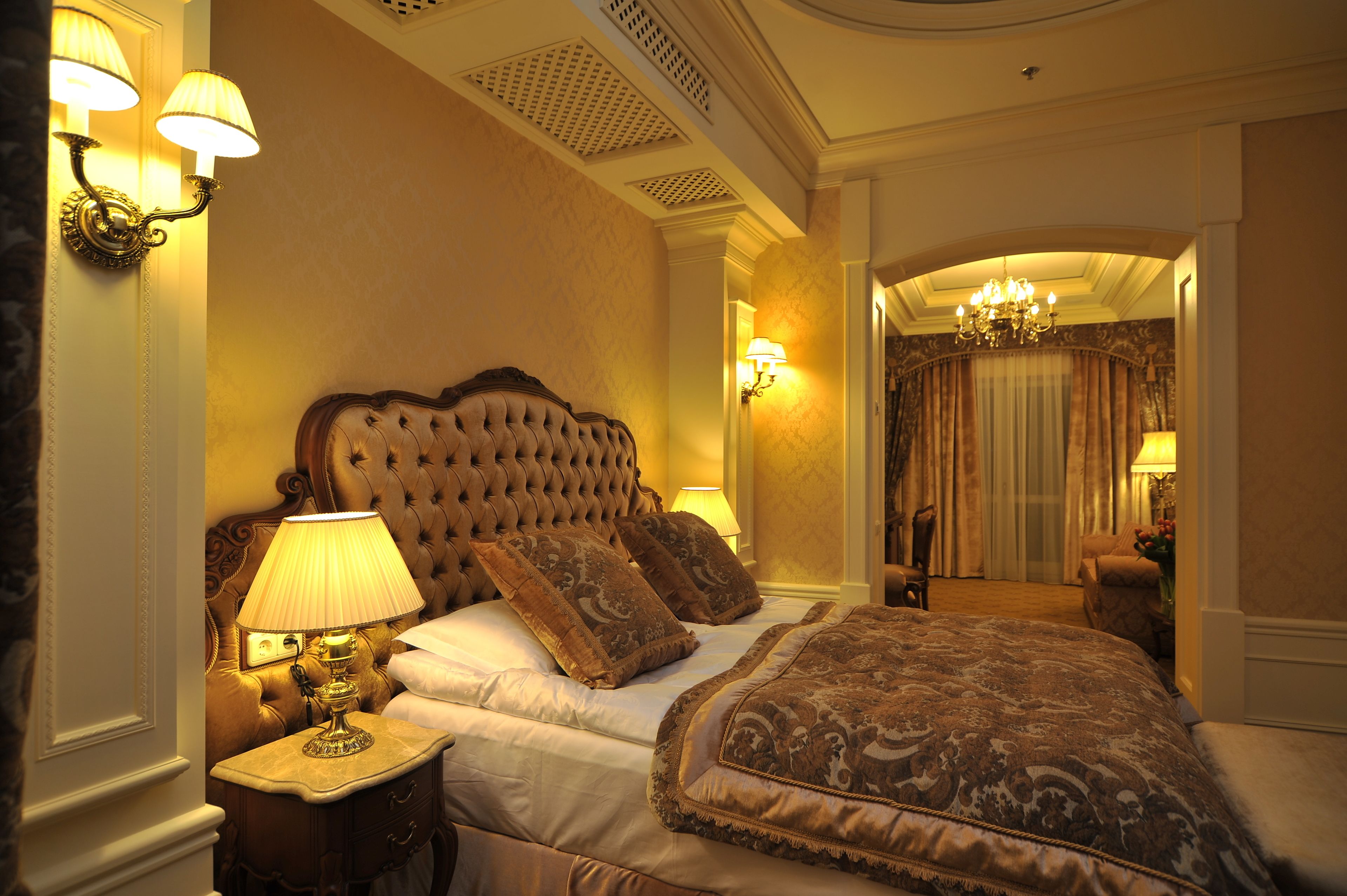 Un dormitorio lujoso con un cabecero capitonado, una cama grande con ropa de cama ornamentada, una mesita de noche con una lámpara y vistas a un pasillo con un candelabro.