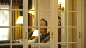 A hotel room with a large window, a bed with a green and blue patterned bedspread, a nightstand with a lamp, and a wall-mounted mirror.