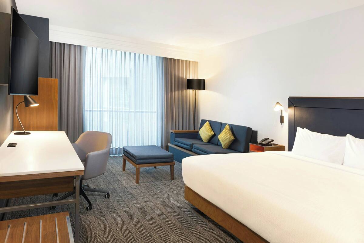 A hotel room with a large bed, a desk with a chair, a sofa, and a TV mounted on the wall.