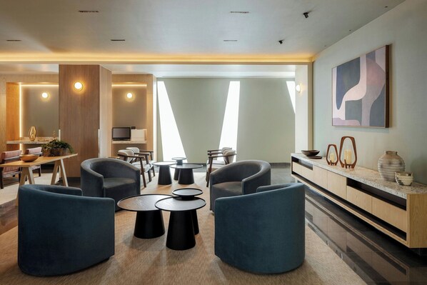 Un moderno vestíbulo de hotel con asientos curvos, mesas redondas, un mostrador de recepción de madera y un gran cuadro abstracto en la pared.