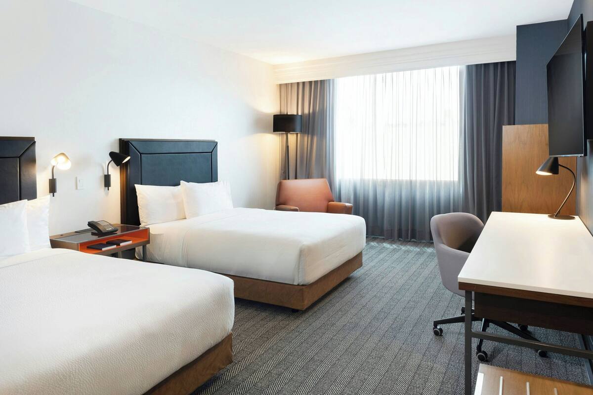 A hotel room with two beds, a desk, a chair, a TV, and a window with curtains.