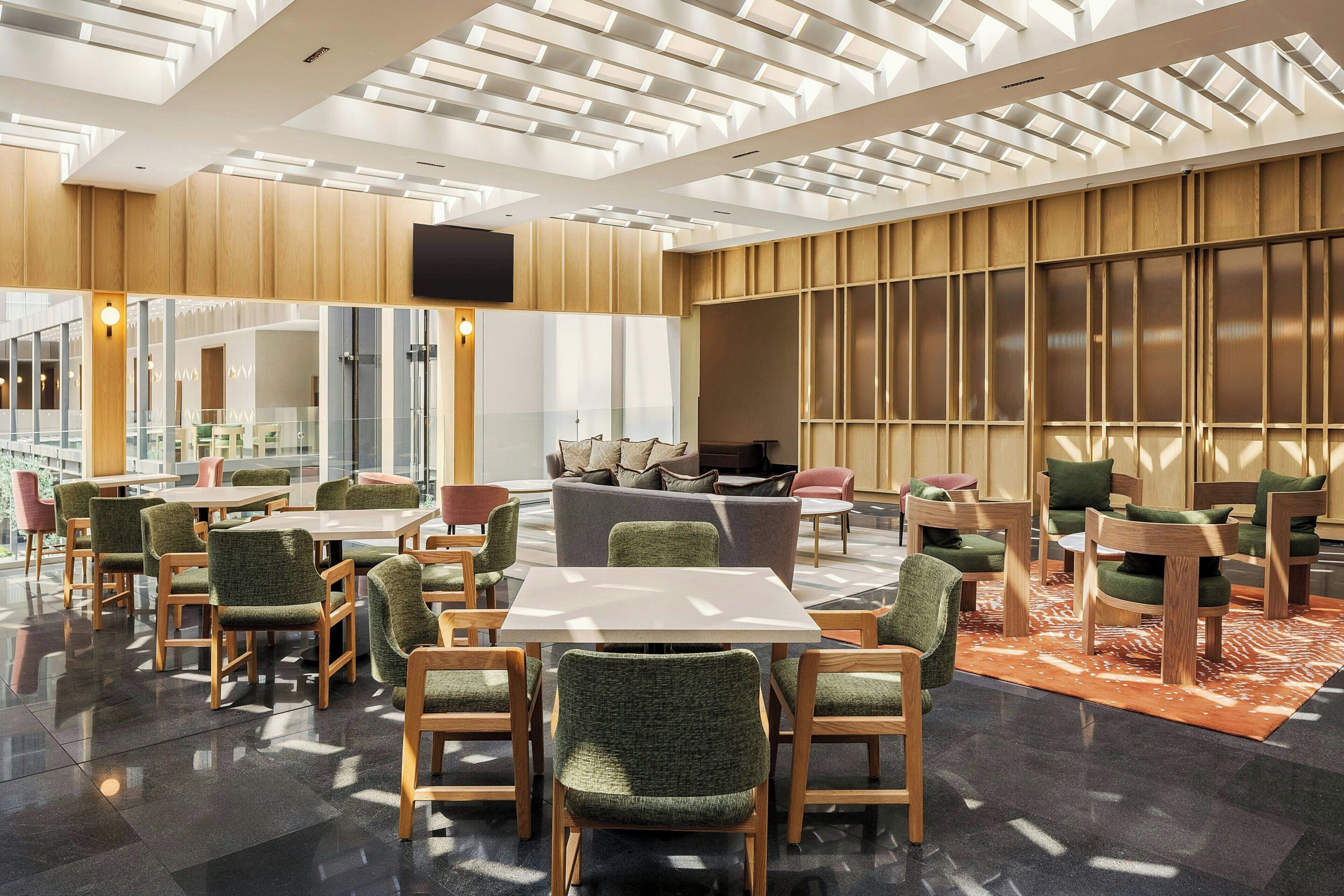 A modern hotel lobby with a central seating area, a flat-screen TV mounted on the wall, and a large window providing natural light.