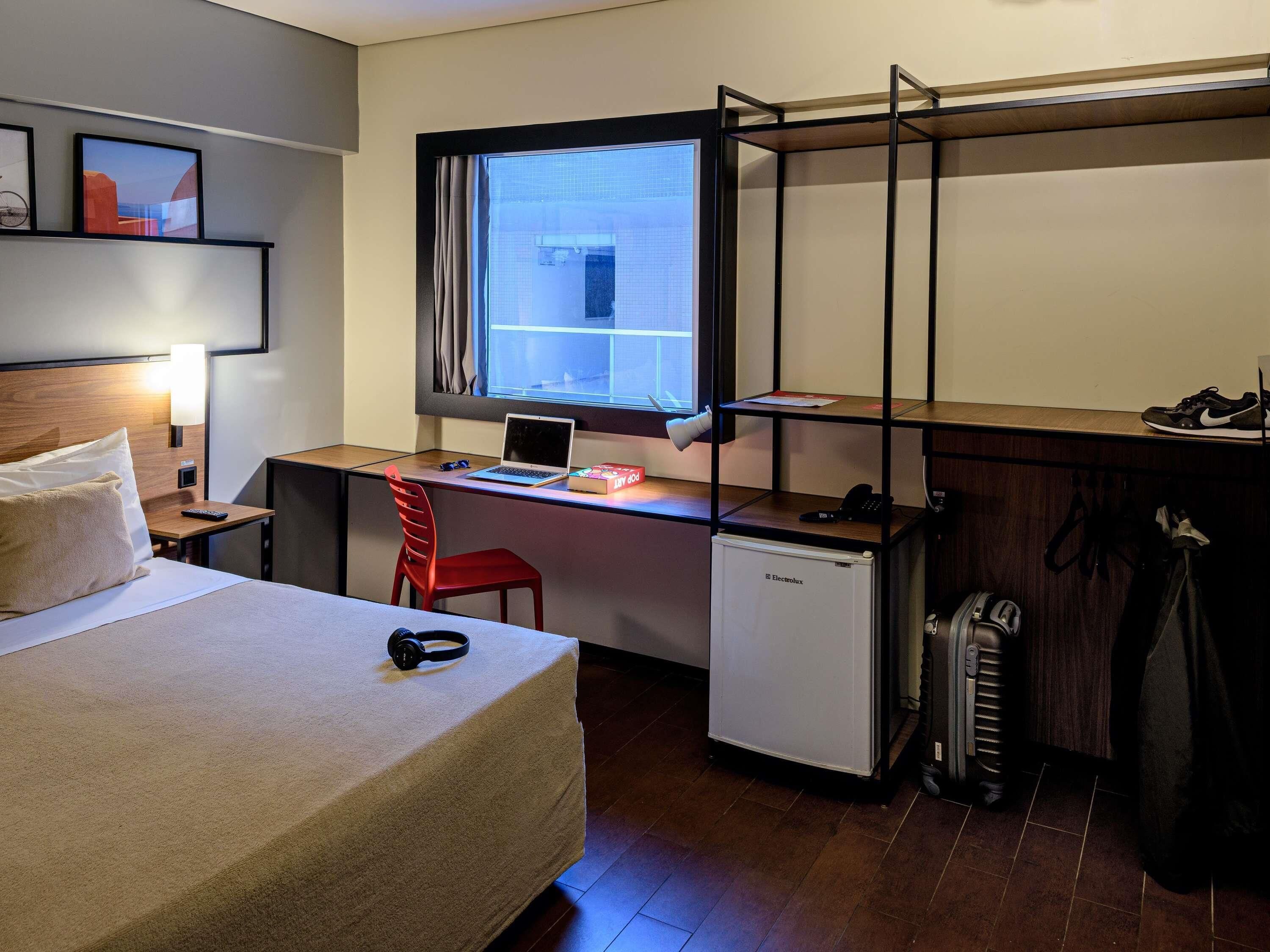 A compact hotel room with a bed, a desk with a laptop, a small refrigerator, a red chair, and a window with curtains.