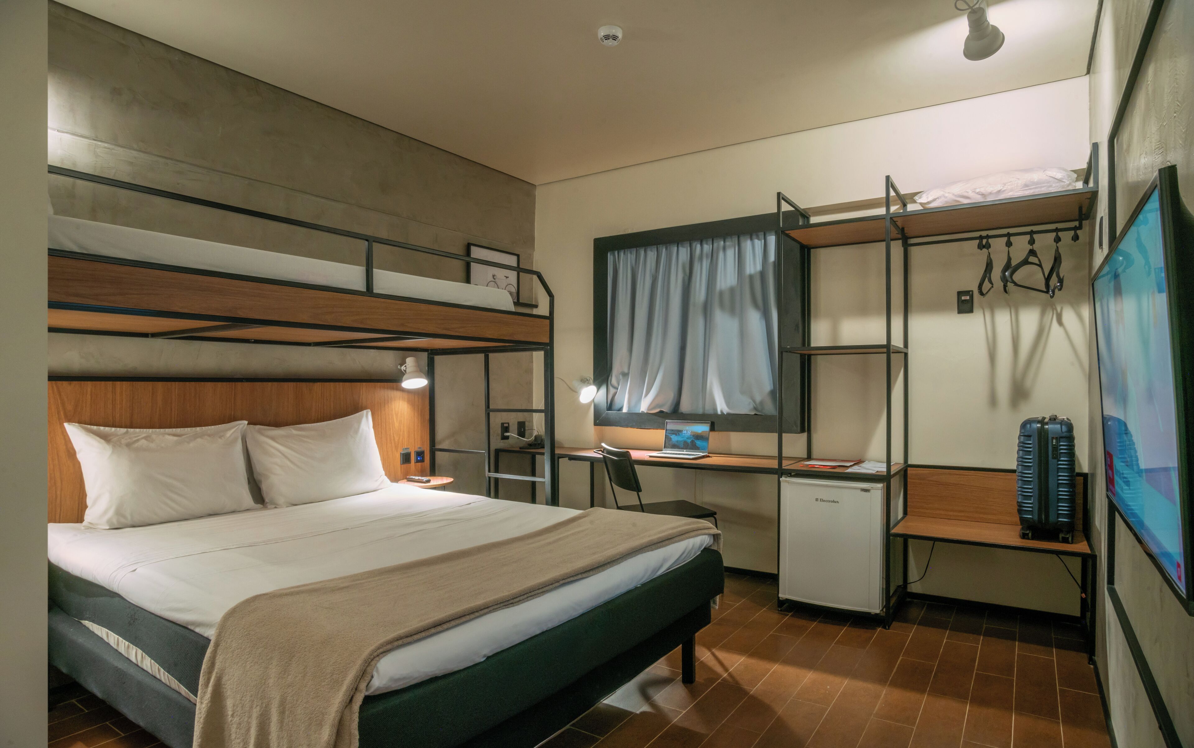 A compact hotel room with bunk beds, a desk with a computer, a small refrigerator, and a TV.