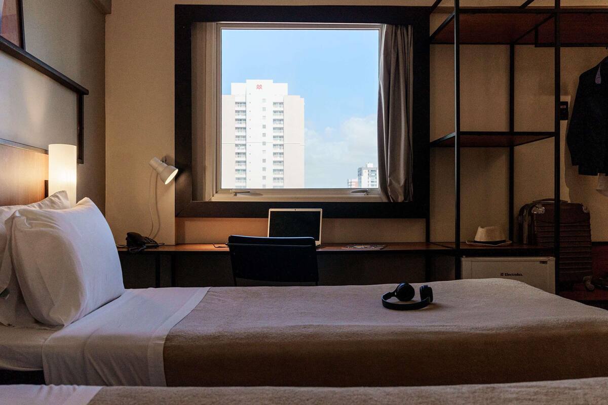 Quarto de hotel com cama, escrivaninha, laptop, janela com vista para a cidade e guarda-roupa.