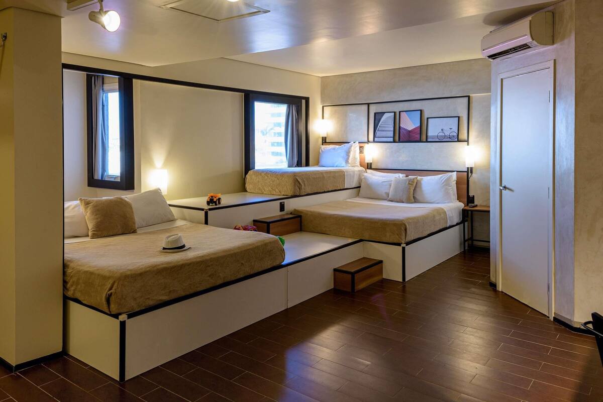 Quarto de hotel com duas camas, uma mesa pequena, luminária, janela e ar condicionado.