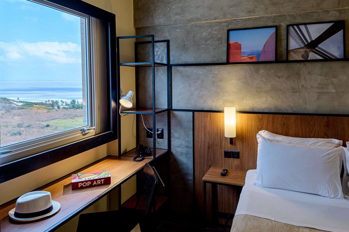 Quarto de hotel com uma cama, uma escrivaninha com abajur, um telefone, um chapéu e um livro intitulado 'POP ART'.