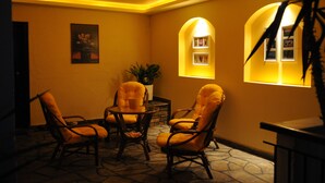 A warmly lit room with four cushioned chairs around a small table, a potted plant, and two illuminated windows. - Hotel Petunia (Sithonia)