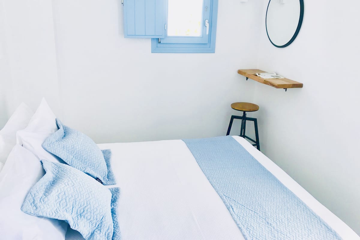 A neatly arranged bedroom with a bed, pillows, a small stool, a shelf, a mirror, and a window with a blue shutter.