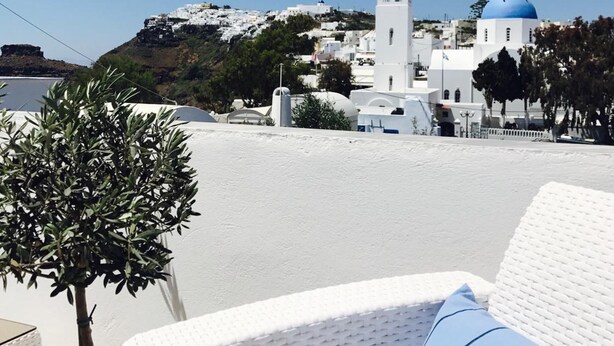Una silla de mimbre blanca con un cojín azul, una pequeña planta en maceta, y vistas de un edificio blanco con cúpulas azules y una torre con campana, todo bajo un cielo azul despejado.