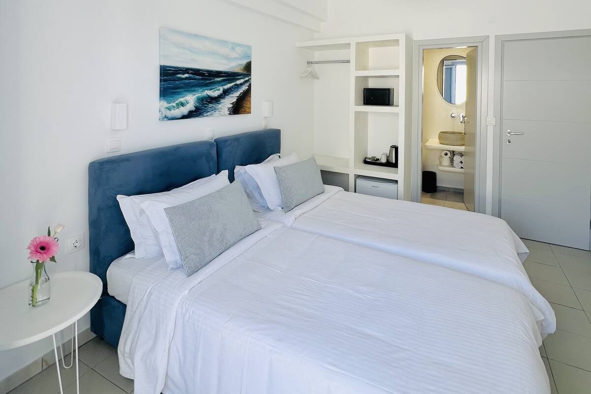 A modern hotel room with a large bed, a bedside table with a vase of flowers, a painting of the sea, and a bathroom visible through an open door.