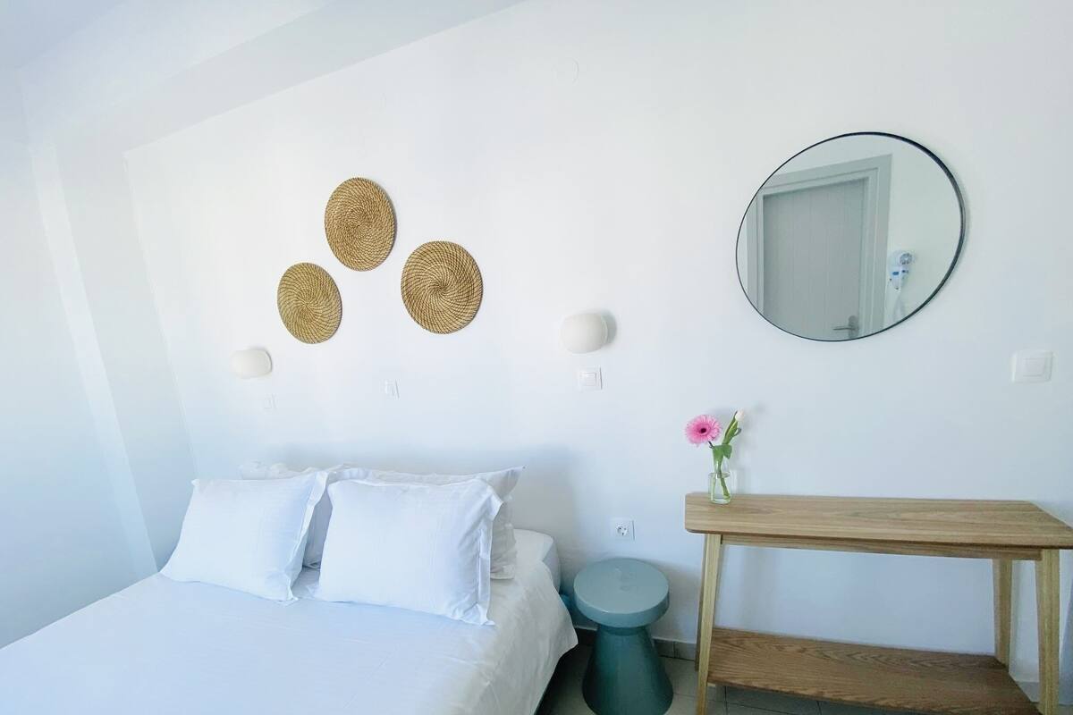 A bedroom with a bed, a round mirror, a wooden bedside table, a small round stool, and decorative woven wall art.