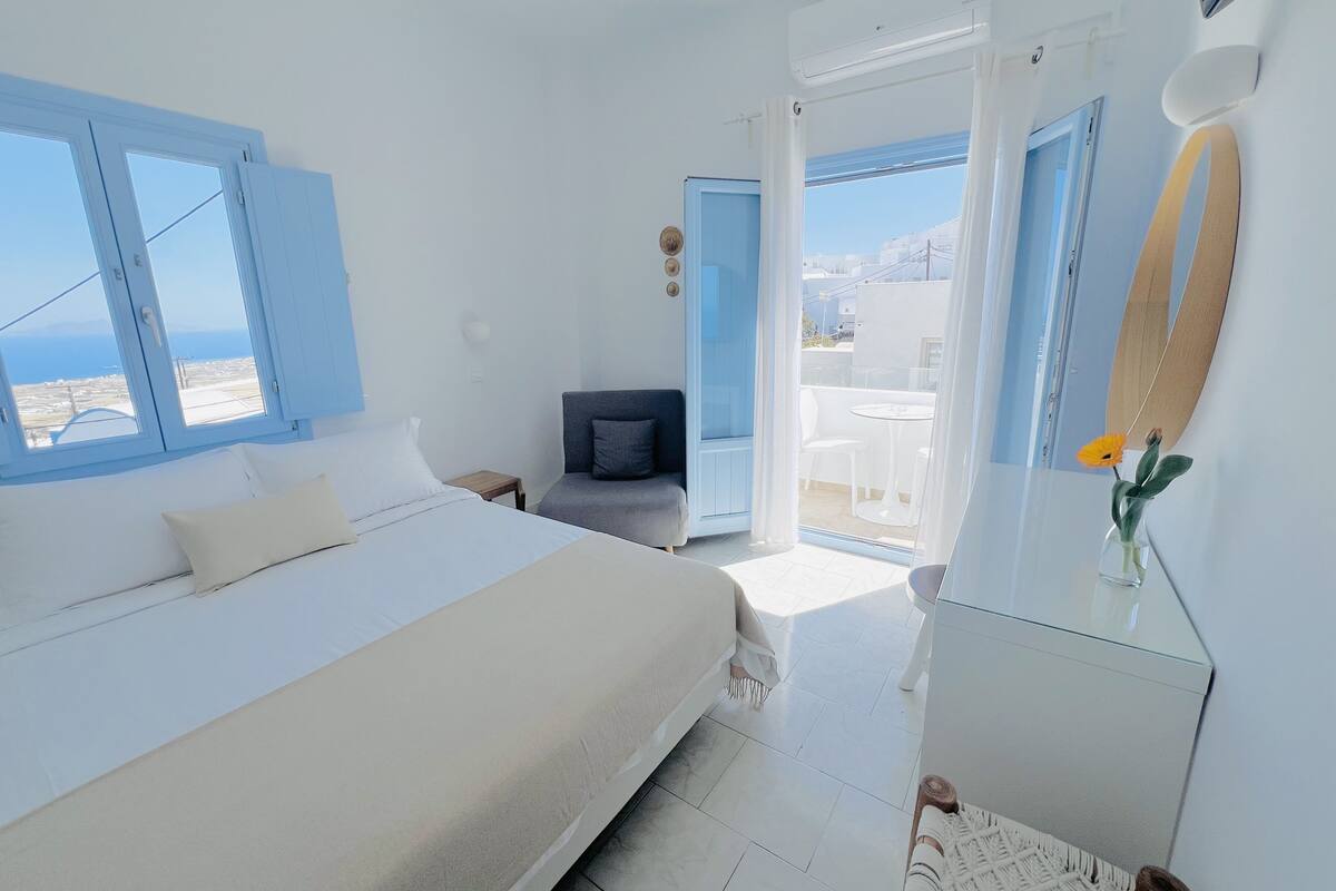 A bedroom with a bed, a small table, a chair, and a view of the outside.