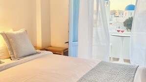 Habitación de hotel con cama, mesita de noche, una pequeña mesa con dos vasos y vistas a edificios de cúpulas azules a través de una ventana con cortinas traslúcidas.