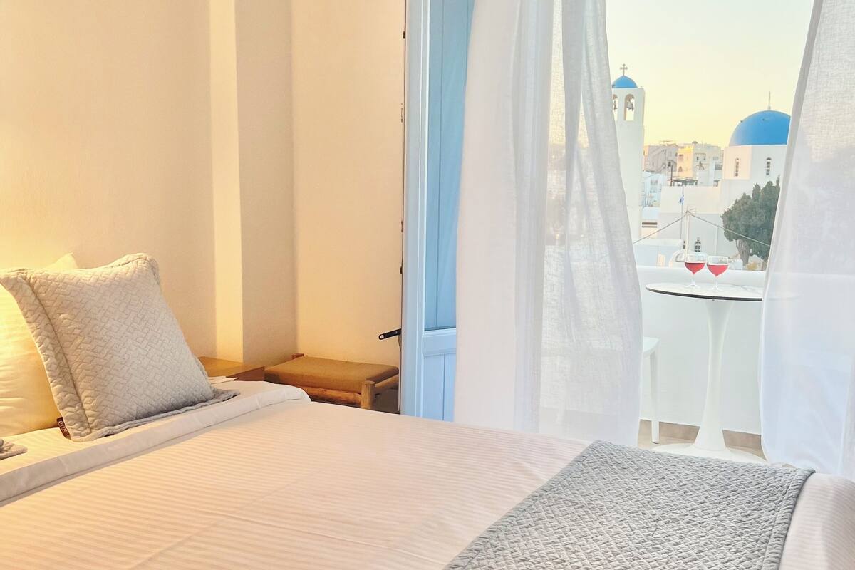 A hotel room with a bed, a bedside table, a small table with two glasses, and a view of blue-domed buildings through a window with sheer curtains.