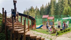 A playground with wooden climbing structures and a slide, surrounded by a fenced area and greenery. - Kamelia (Smolyan)