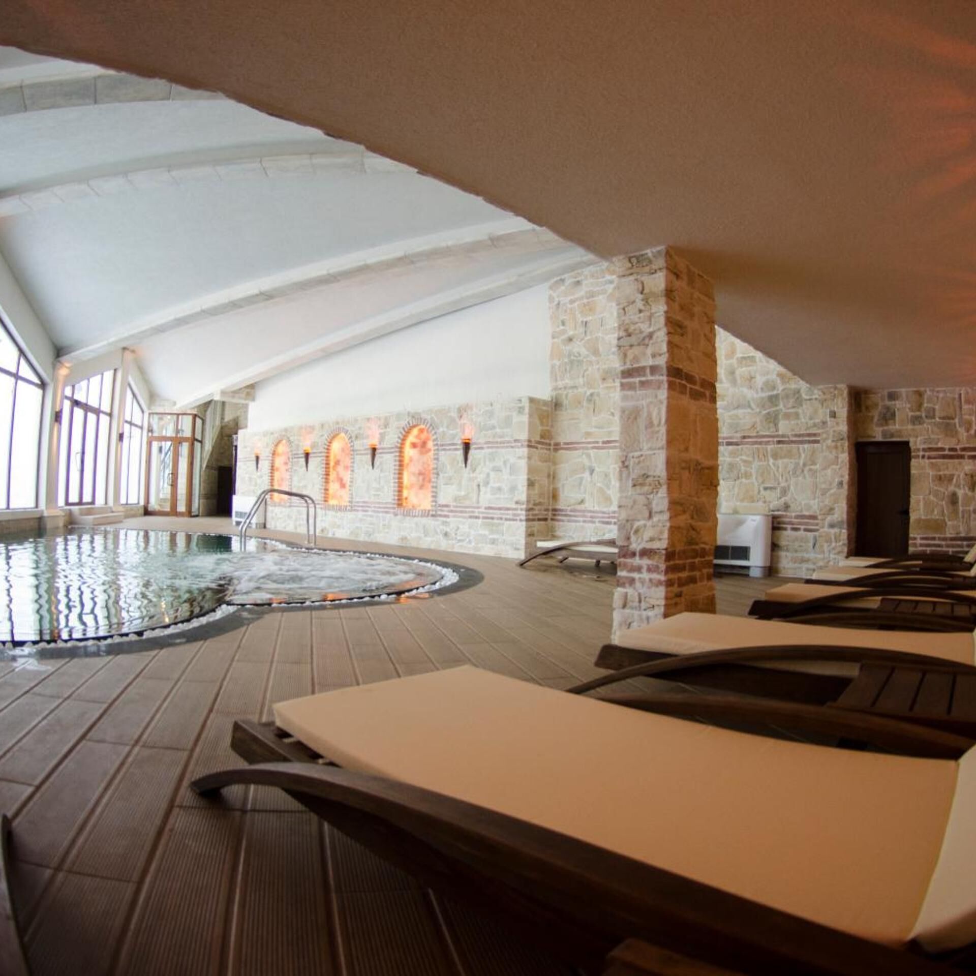 An indoor swimming pool with lounge chairs, stone walls, and large windows.