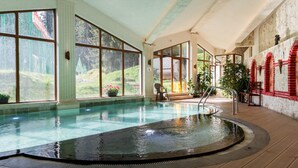 A large indoor swimming pool with clear glass walls, wooden deck, and a stone wall with red accents. - Kamelia (Smolyan)