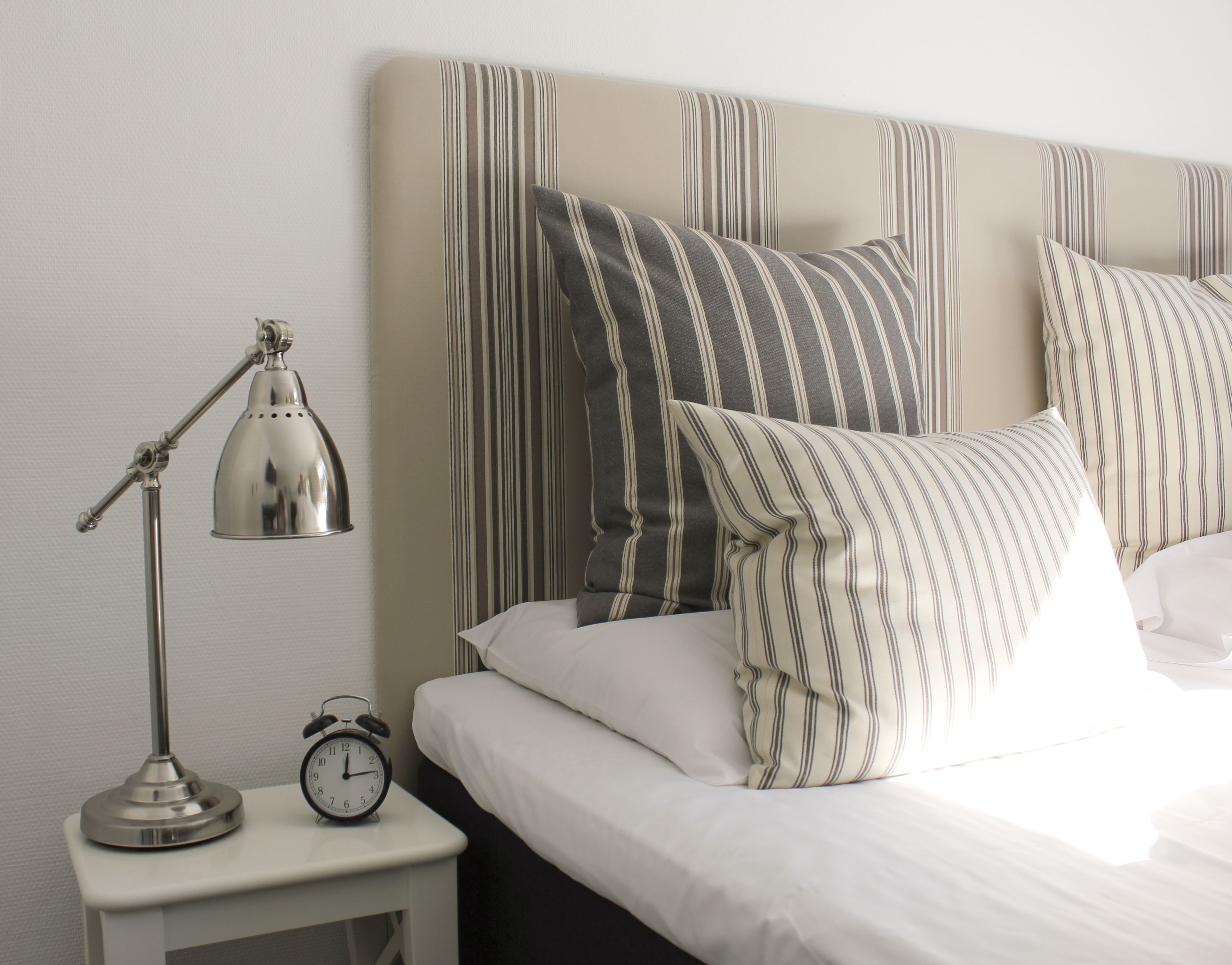 A neatly made bed with striped pillows and a bedside table with a lamp and an alarm clock.