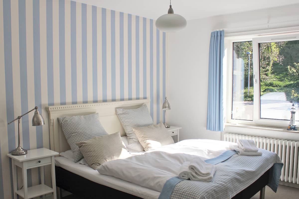 Una cama bien hecha con almohadas, una mesita de noche con una lámpara y una ventana con cortinas.