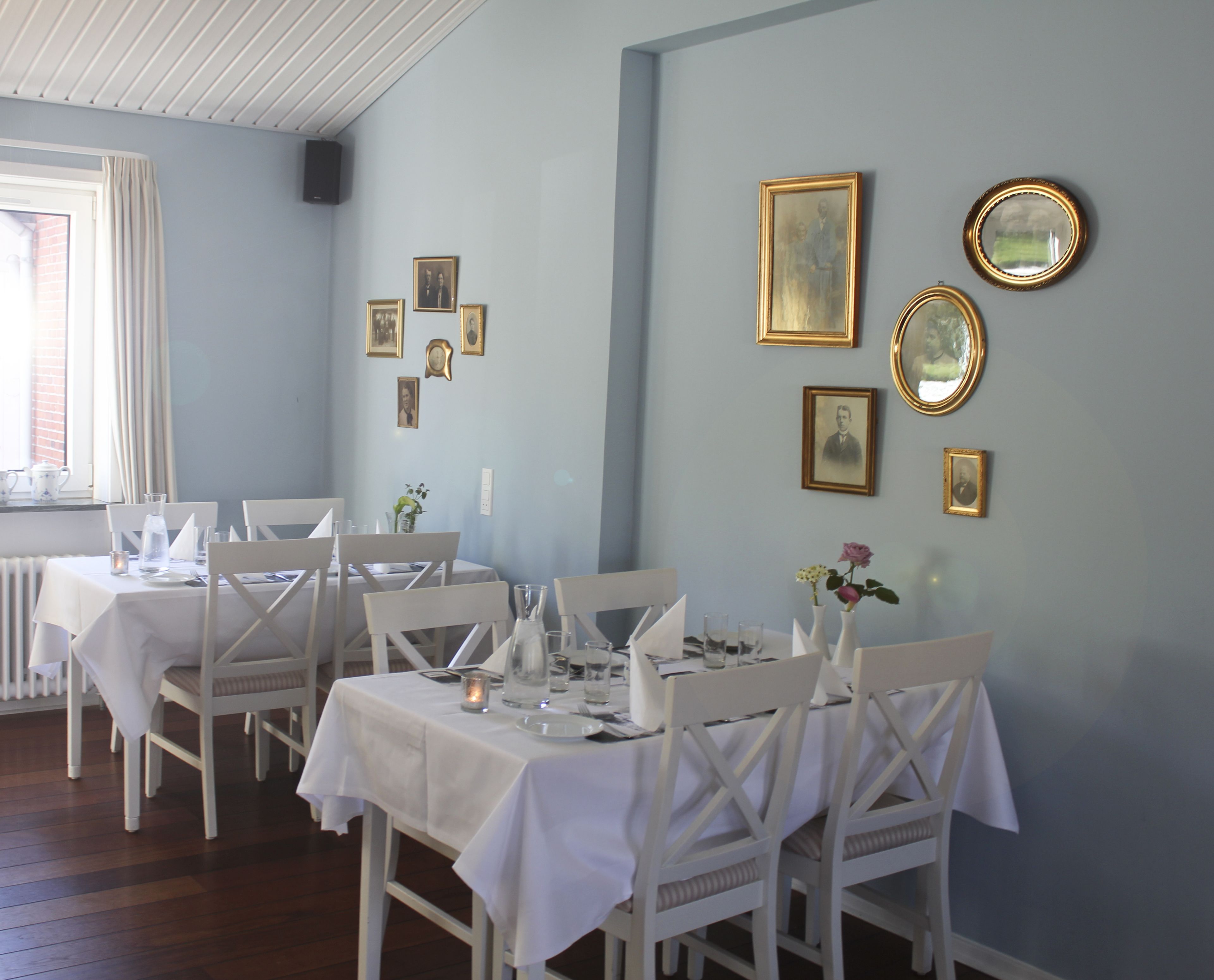 Un espace repas avec des tables et des chaises blanches, des tableaux encadrés au mur et une fenêtre avec des rideaux.
