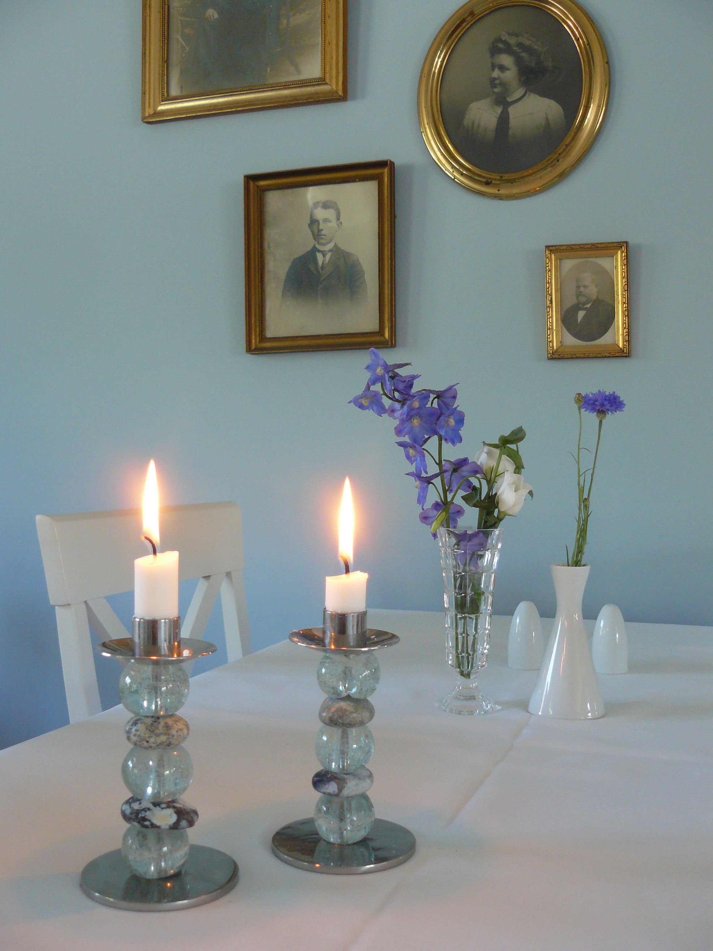 A room with a table set for a meal, featuring two lit candles, a vase with flowers, and framed pictures on the wall.